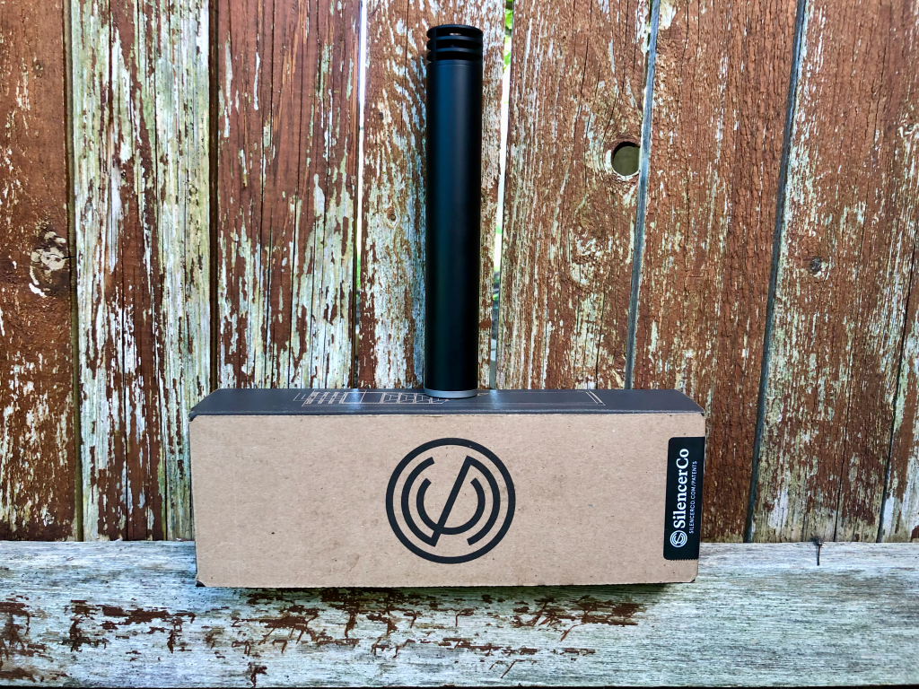 A black SilencerCo Omega 300 suppressor standing on its SilencerCo box against a weathered wood fence