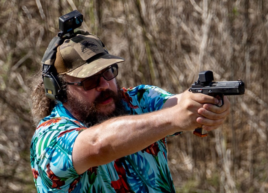 Uncle Zo in a colorful Hawaiian shirt aiming a pistol at a shooting range, wearing ear protection and a tactical hat at the 2023 USPSA Primary Arms Space City Challenge.